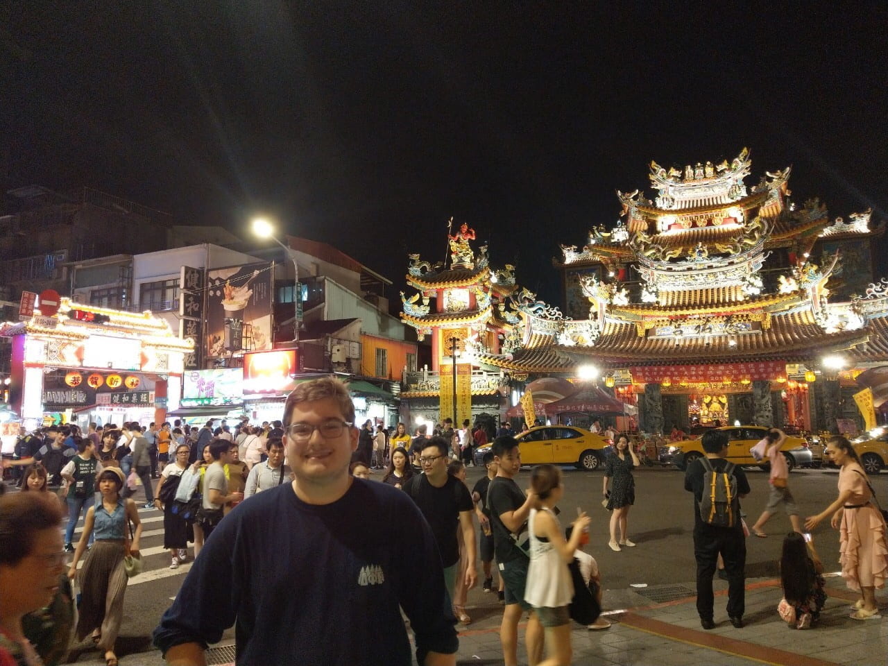 Sigur language fellow poses in front of a large gate at a busy intersection at night in Taipei in front of a busy street with the Raohe Night market and temple gate behind him