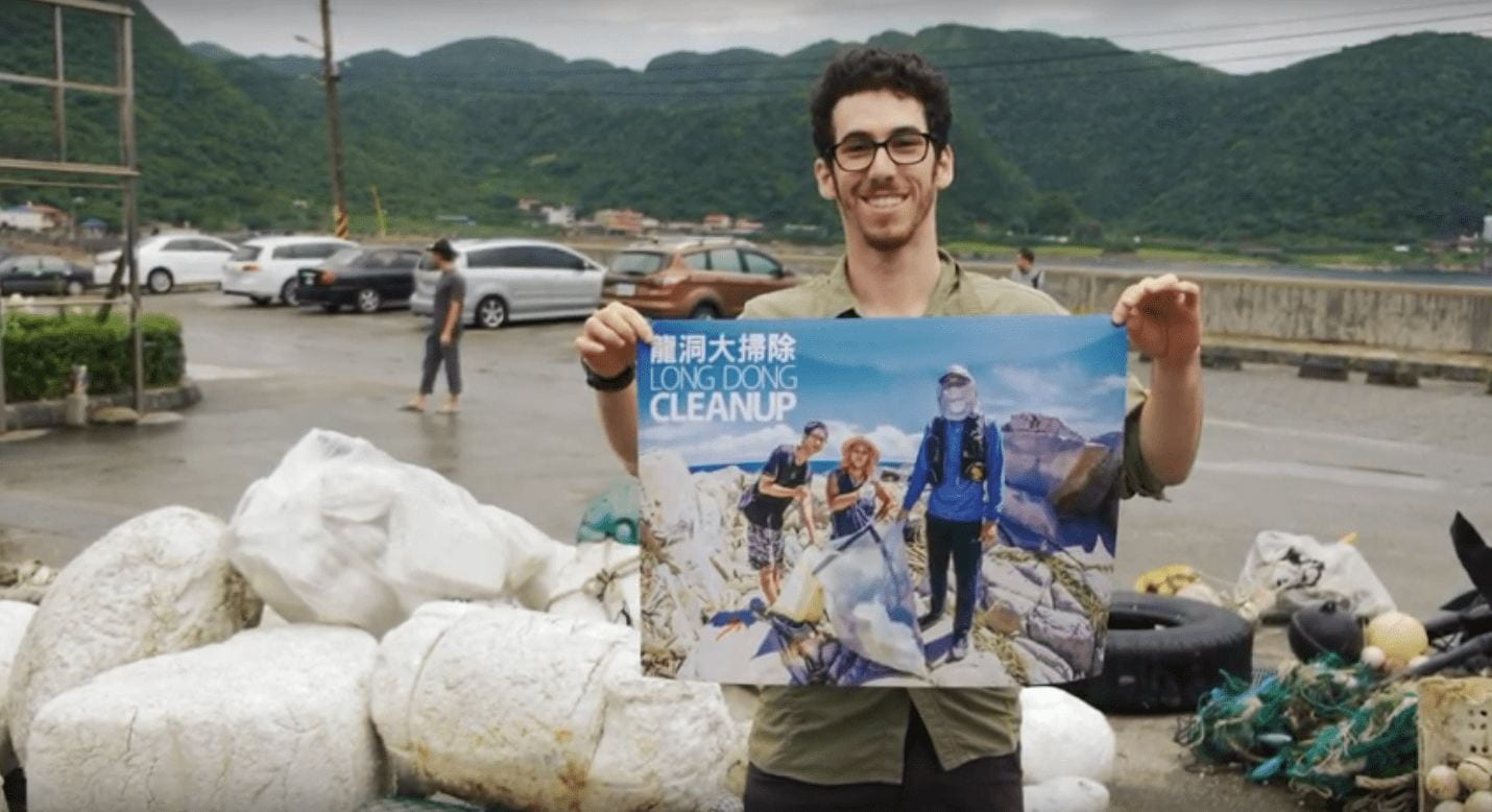 Jason Shor at the beach standing next to a pile of trash reclaimed from the ocean holding up a poster advertising the local efforts to clean up the beach, Poster Text: Long Ding Clean Up